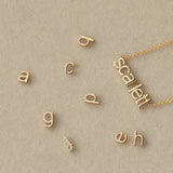 A Petite Nameplate Necklace in Scarlett with several Petite Letter Charms, all in yellow gold, against a light beige background. 