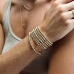 Close-up of a woman’s arm wearing multiple Statement Bubble Bracelets, gold necklaces and a Link Ring, in a light sage green top.