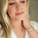 A woman wearing a Custom Gold Necklace - 3 Letters, a Classic Bead Chain with a Princess Diamond Pendant, an Element Short Link Chain, Element Long Link Chain, Rolo Bracelet and Diamond Cut Bracelet, all in yellow gold, with blonde hair and a white top against a blurred background.
