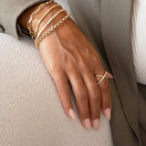 A close-up of a woman’s hand on a cream surface wearing stacked yellow gold bracelets: a Bold Link Bracelet, an Element Short Link Bracelet, a Love Set Match Bracelet, a Down the Line Bracelet and a Rolo Bracelet, and a Criss-Cross Diamond Daze Ring, with a sage green jacket and white top.