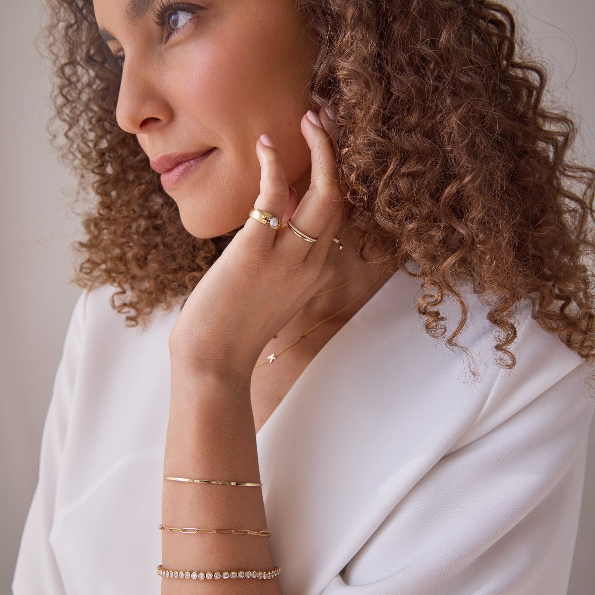 A woman with curly hair resting her hand on her face, wearing an Element Long Link Bracelet, a Love Set Match Bracelet, an Off the Cuff Bracelet, a Perla Pinky Ring, a Delicate Band, a Pavé Diamond Ring and a Custom Gold Necklace with a letter K charm, all in yellow gold, styled with a white long sleeve top.