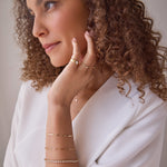 A woman with curly hair resting her hand on her face, wearing an Element Long Link Bracelet, a Love Set Match Bracelet, an Off the Cuff Bracelet, a Perla Pinky Ring, a Delicate Band, a Pavé Diamond Ring and a Custom Gold Necklace with a letter K charm, all in yellow gold, styled with a white long sleeve top.