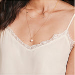 A close-up of a woman’s chest wearing a cream lace top and three yellow gold necklaces: a Perla Necklace, an Angel Wing Charm Necklace and a Custom Gold Necklace with four charms (three birthstones and a Star of David).