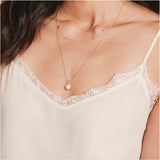 A close-up of a woman’s chest wearing a cream lace top and three yellow gold necklaces: a Perla Necklace, an Angel Wing Charm Necklace and a Custom Gold Necklace with four charms (three birthstones and a Star of David).
