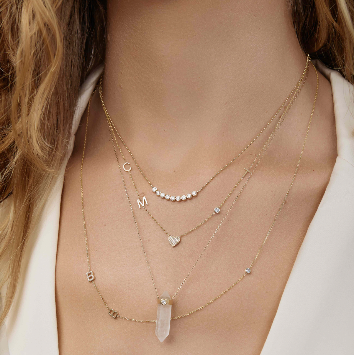 A close-up of a woman’s neck wearing four yellow gold necklaces: two Custom Gold Necklaces (one with three charms and the other with four charms), the Clarity Retreat Necklace and the Diamond Arc Layering Necklace, with a cream top.