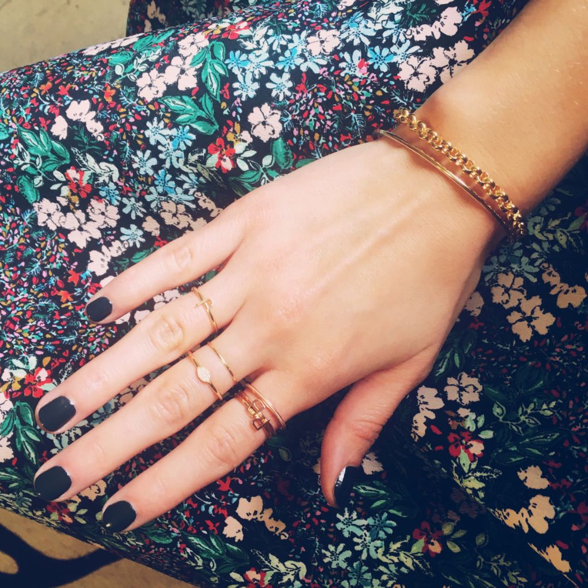 A hand wearing yellow and rose gold jewellery, including a Simple Bangle, a Rolo Bracelet, three 14k Gold Letter Rings (J, R and B), two Delicate Bands and a 14k Gold Pavé Petal Ring with black nail polish against a floral fabric background.