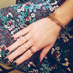 A hand wearing yellow and rose gold jewellery, including a Simple Bangle, a Rolo Bracelet, three 14k Gold Letter Rings (J, R and B), two Delicate Bands and a 14k Gold Pavé Petal Ring with black nail polish against a floral fabric background.