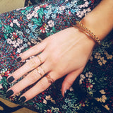 A hand wearing yellow and rose gold jewellery, including a Simple Bangle, a Rolo Bracelet, three 14k Gold Letter Rings (J, R and B), two Delicate Bands and a 14k Gold Pavé Petal Ring with black nail polish against a floral fabric background.