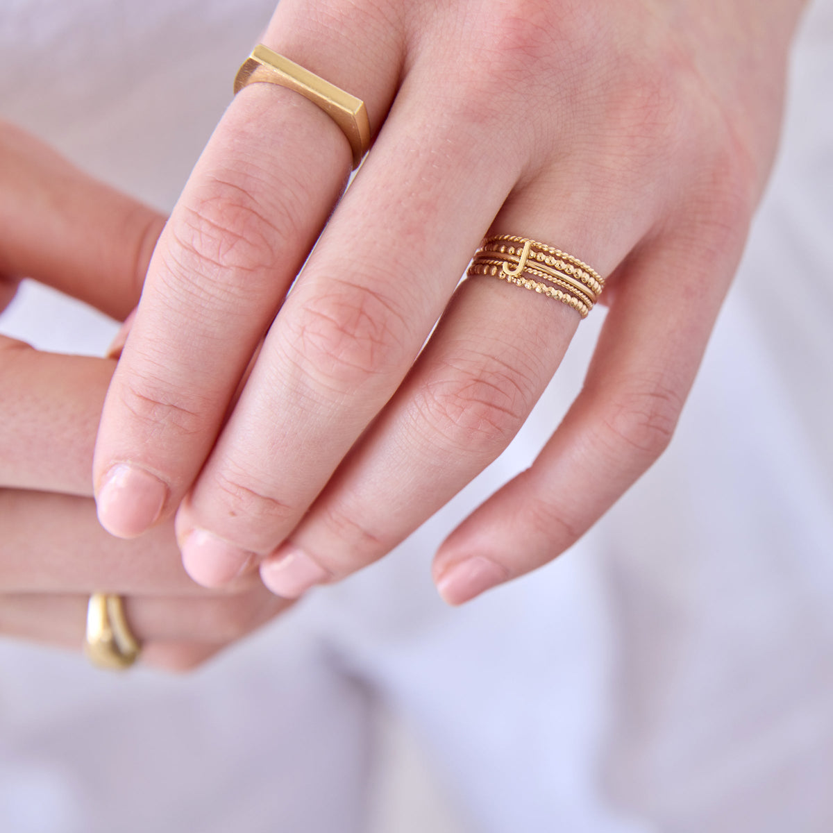 Close-up of hands wearing Delicate Trio Stacking Rings, a Geo Stacking Ring Square and a 14k gold Letter Ring with J, all in yellow gold on a light background. 