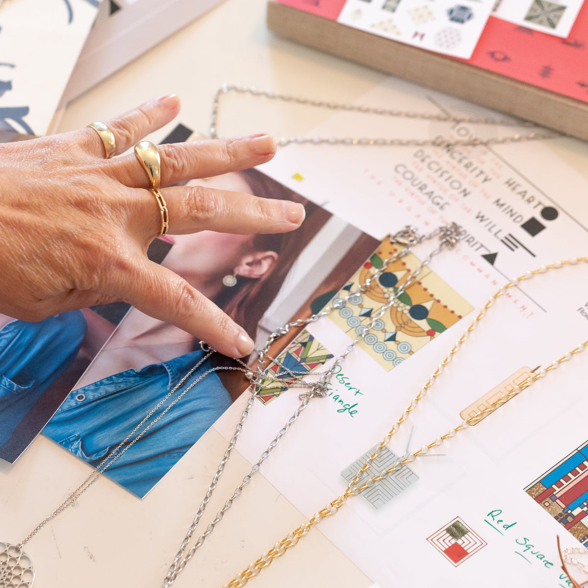 A hand wearing a Pinky Ring, Paris Ring and Link Ring in yellow gold, resting on a table with Virtues Pendants and a Skylight Necklace laid out on a cream surface with photos and papers.