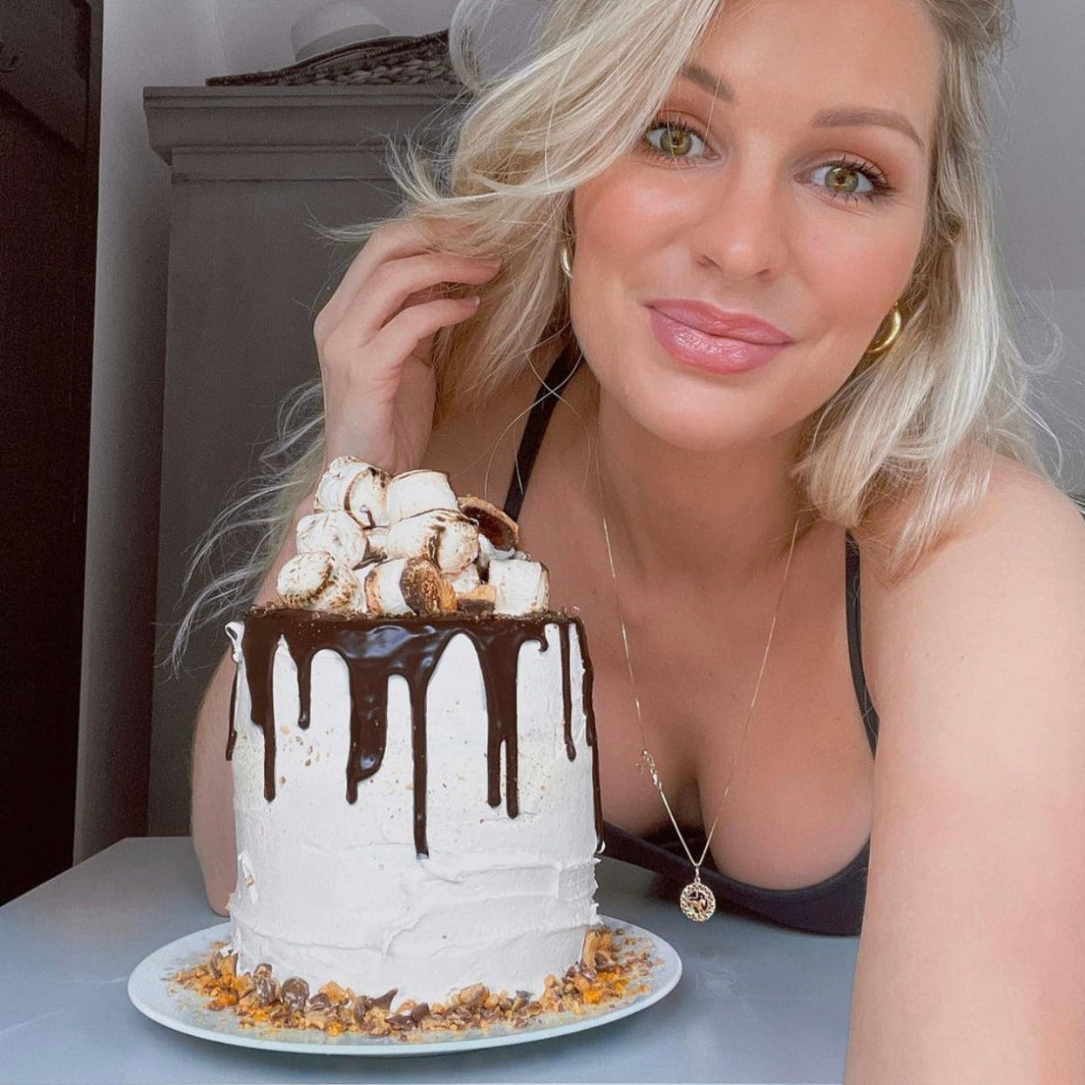 Olivia Newman Young wearing a Zodiac Necklace - Taurus with gold hoops, a black singlet top posting next to a cake with chocolate drizzle and marshmallows. 