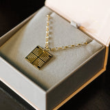A Virtues Pendant - Square in yellow gold in a Maya Brenner light grey and pink jewellery box against a black background. 