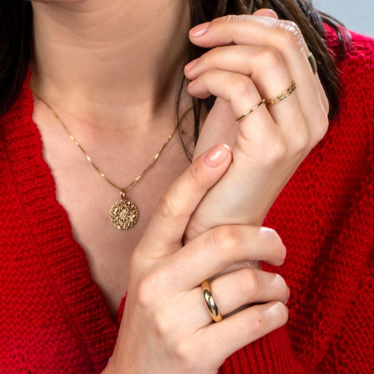 Close-up of a brunette woman wearing a Zodiac Necklace - Cancer, a Link Ring, a Tube Ring,and a Delicate Band, all in yellow gold, with a red knit against a light grey background.