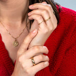 Close-up of a brunette woman wearing a Zodiac Necklace - Cancer, a Link Ring, a Tube Ring,and a Delicate Band, all in yellow gold, with a red knit against a light grey background.