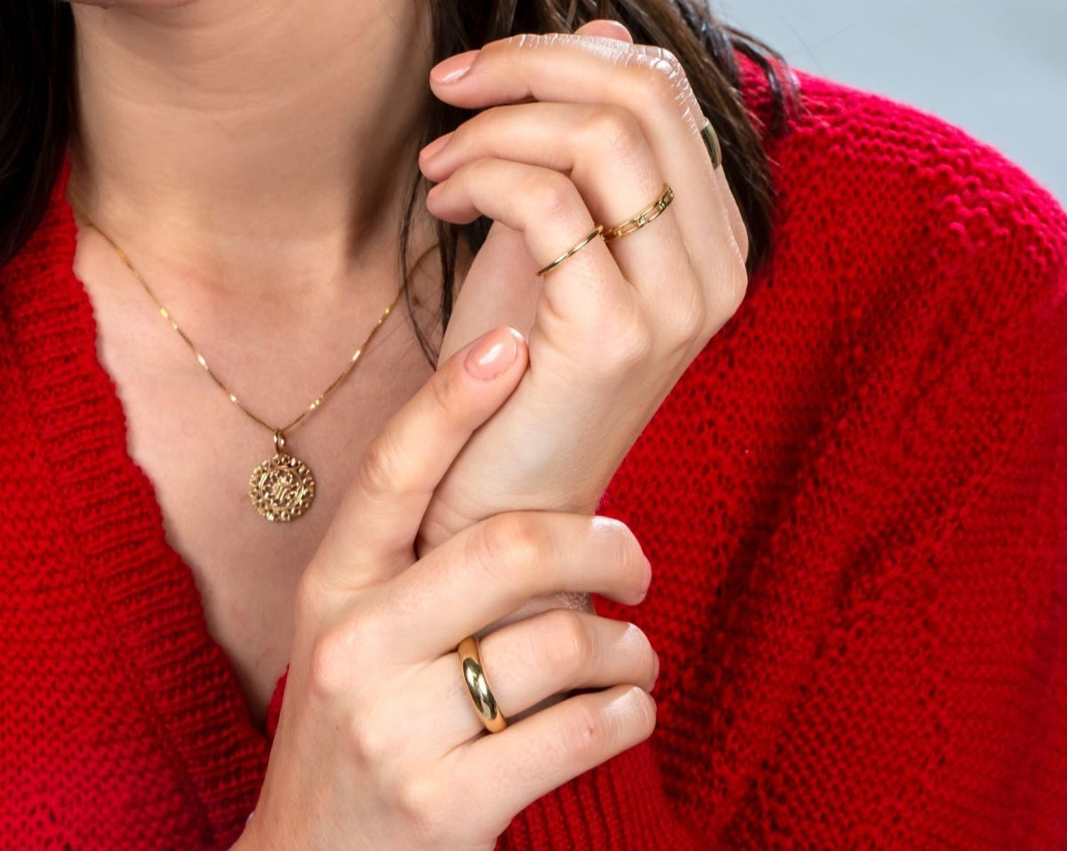 Close-up of a brunette woman wearing a Zodiac Necklace - Cancer, a Link Ring, a Tube Ring,and a Delicate Band, all in yellow gold, with a red knit against a light grey background.