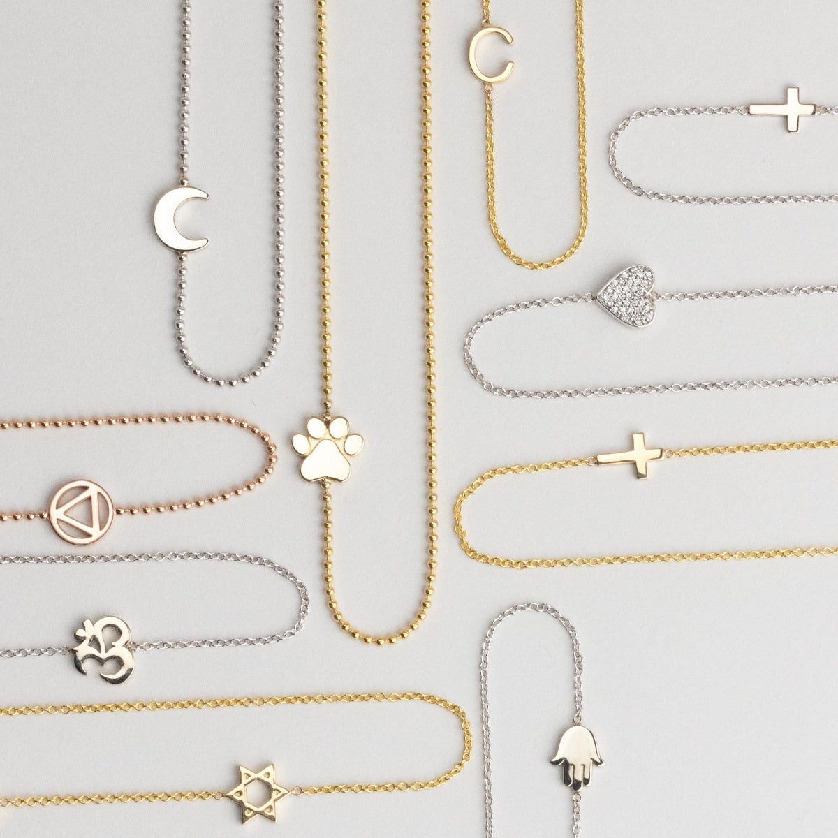 Several 14k Gold Asymmetrical Charm Necklaces in yellow, white and rose gold in both bead and cable chains (moon, paw, recovery, om, star of david, C, cross x 2 and a pavé heart) on a light grey background. 