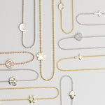 Several 14k Gold Asymmetrical Charm Necklaces in yellow, white and rose gold in both bead and cable chains (moon, paw, recovery, om, star of david, C, cross x 2 and a pavé heart) on a light grey background. 
