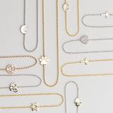 Several 14k Gold Asymmetrical Charm Necklaces in yellow, white and rose gold in both bead and cable chains (moon, paw, recovery, om, star of david, C, cross x 2 and a pavé heart) on a light grey background. 