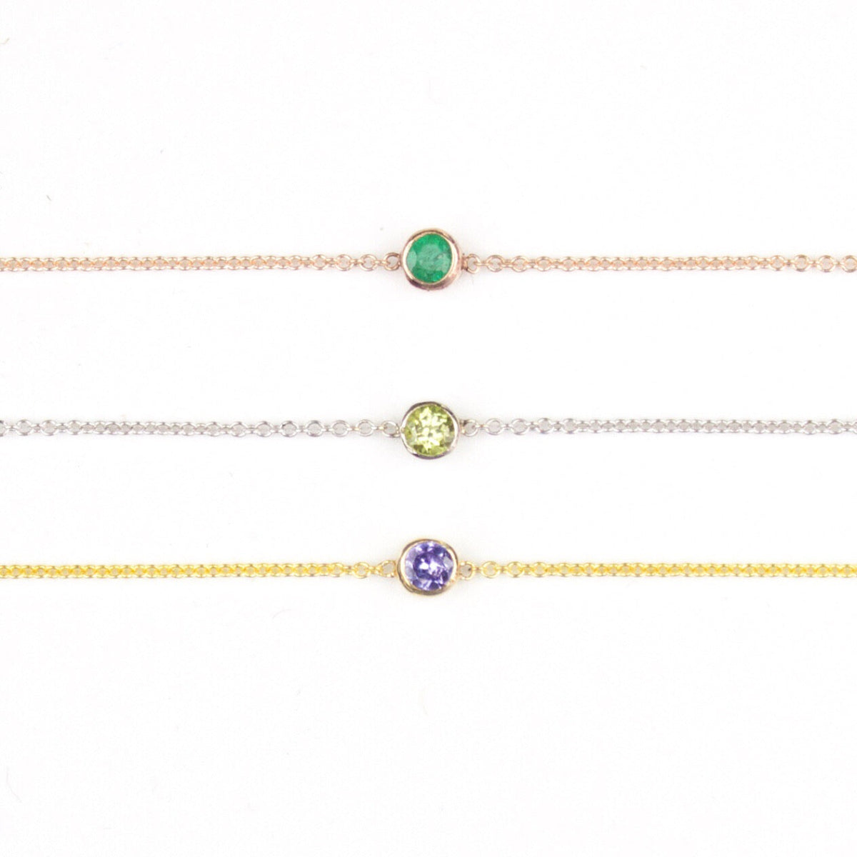 Three 14K Gold Asymmetrical Birthstone Necklaces - Emerald (May) in rose gold, Peridot (August) in white gold and Tanzanite (December) in yellow gold on a white background. 