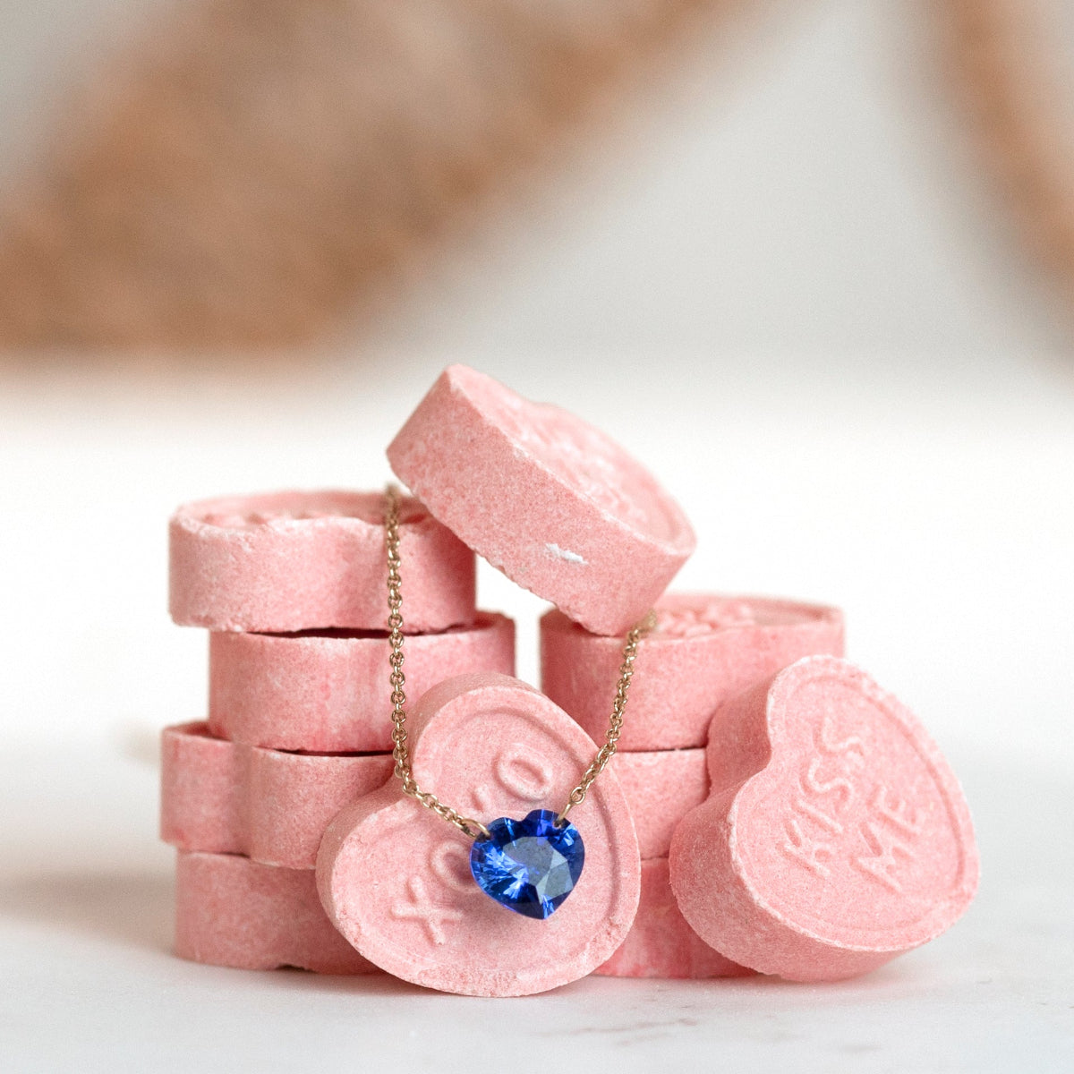 The Sweetheart Necklace - Blue Sapphire laid on top of pink heart-shaped candy with a blurred background. 