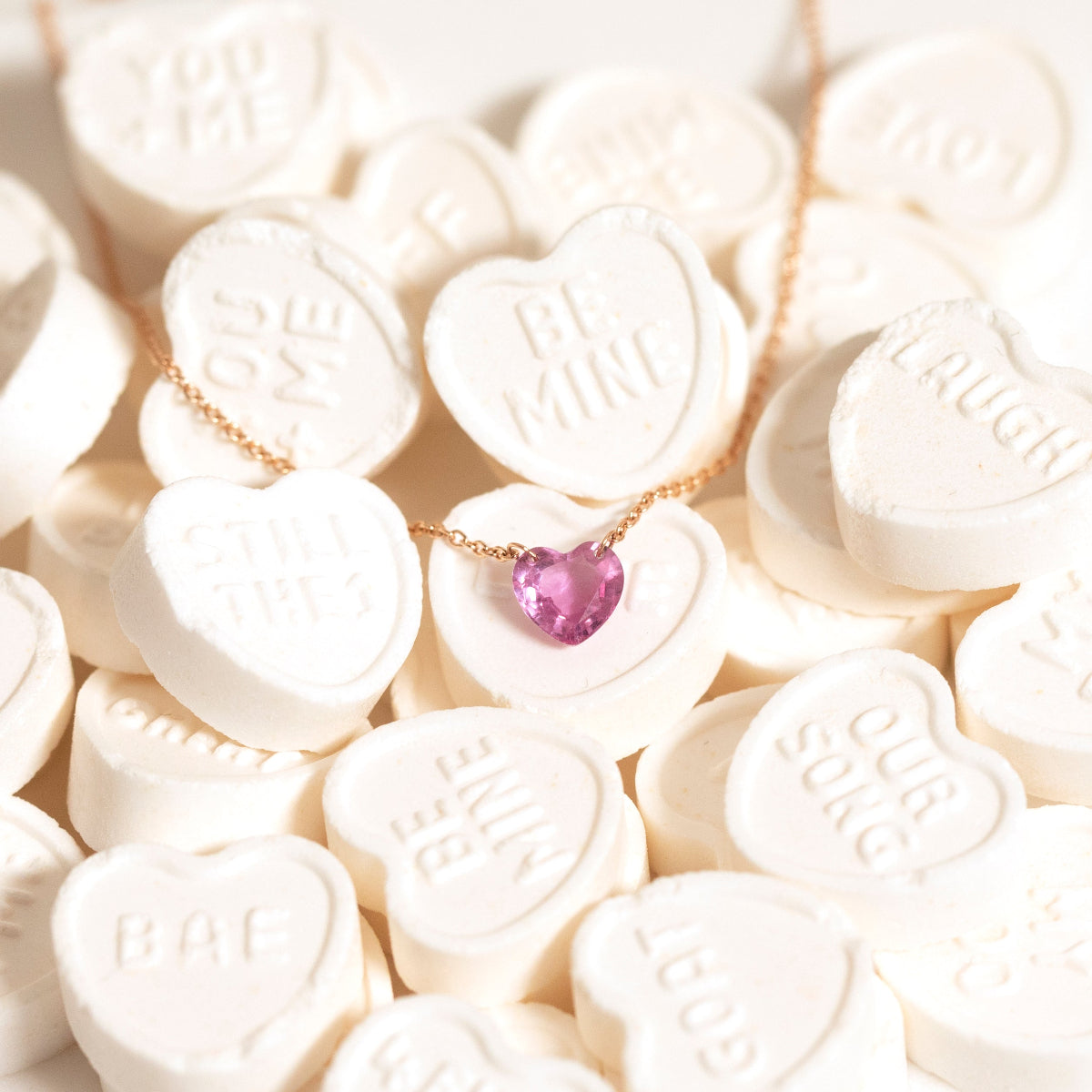 The Sweetheart Necklace - Pink Sapphire laid on top of white heart-shaped candy. 