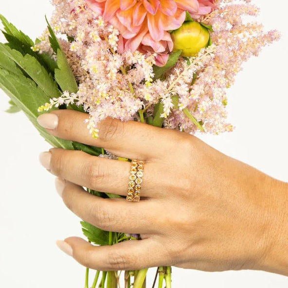 A hand wearing two The Daisy Bouquet Rings holding a bunch of flowers against a light grey background.