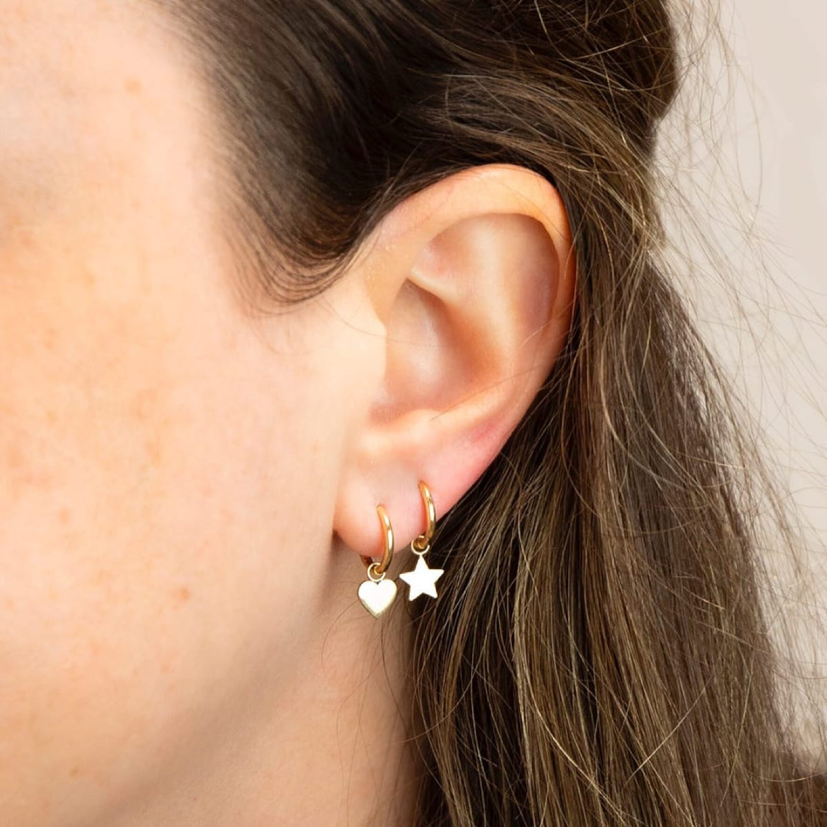 Close-up of Charmed I'm Sure Hoops in Heart and Star shown on a brunette woman's ear against a light grey background. 