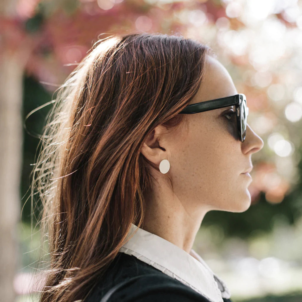 A woman wearing a Large Solid Eclipse Earring, looking to the side, with brown hair, black sunglasses and a black top, outdoors against a softly blurred background.