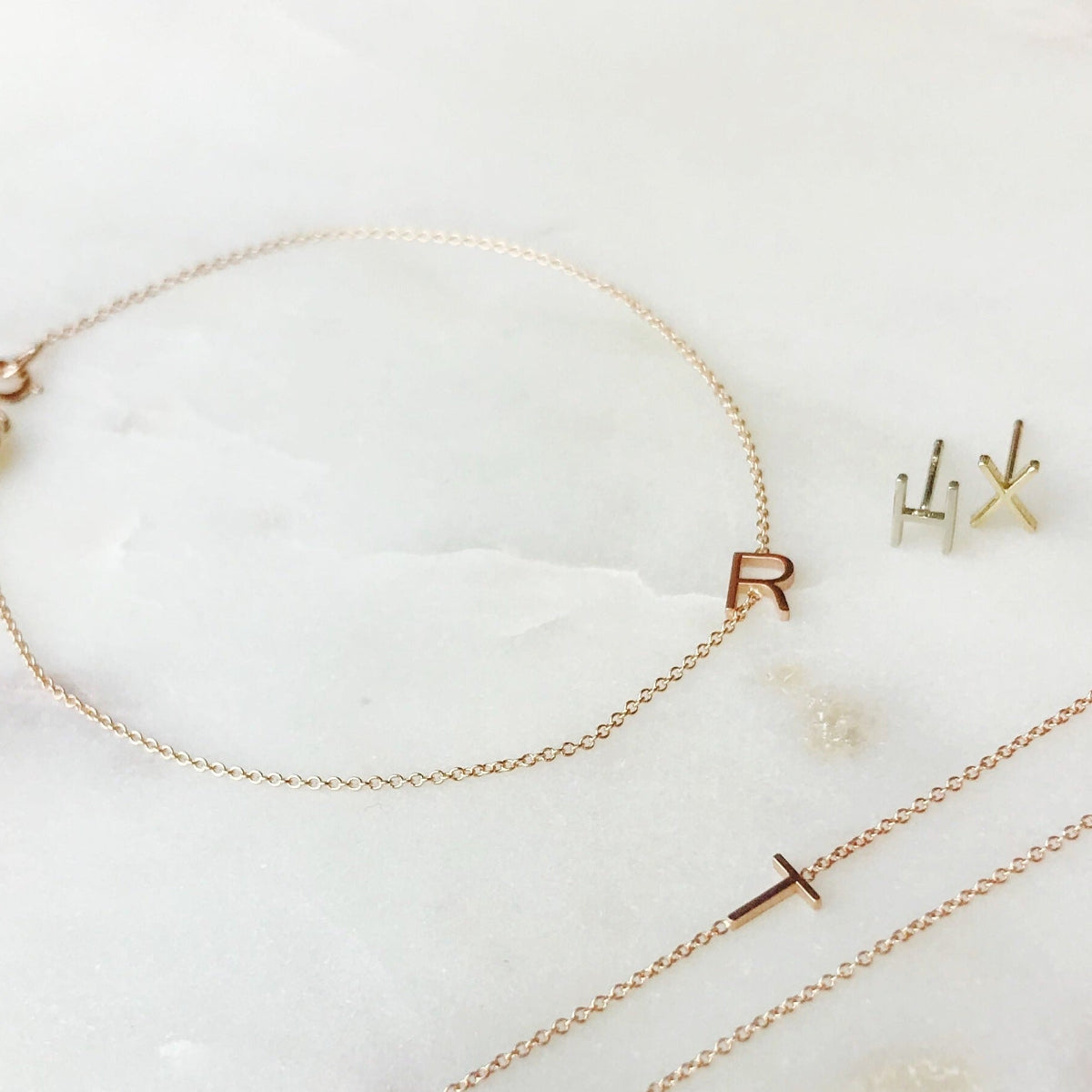 A Custom Gold Bracelet - 1 Letter with the letter R, two 14k Gold Letter Studs in H and X and a Custom Gold Necklace - 1 Letter with the letter T against a light marble background.