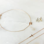 A Custom Gold Bracelet - 1 Letter with the letter R, two 14k Gold Letter Studs in H and X and a Custom Gold Necklace - 1 Letter with the letter T against a light marble background.