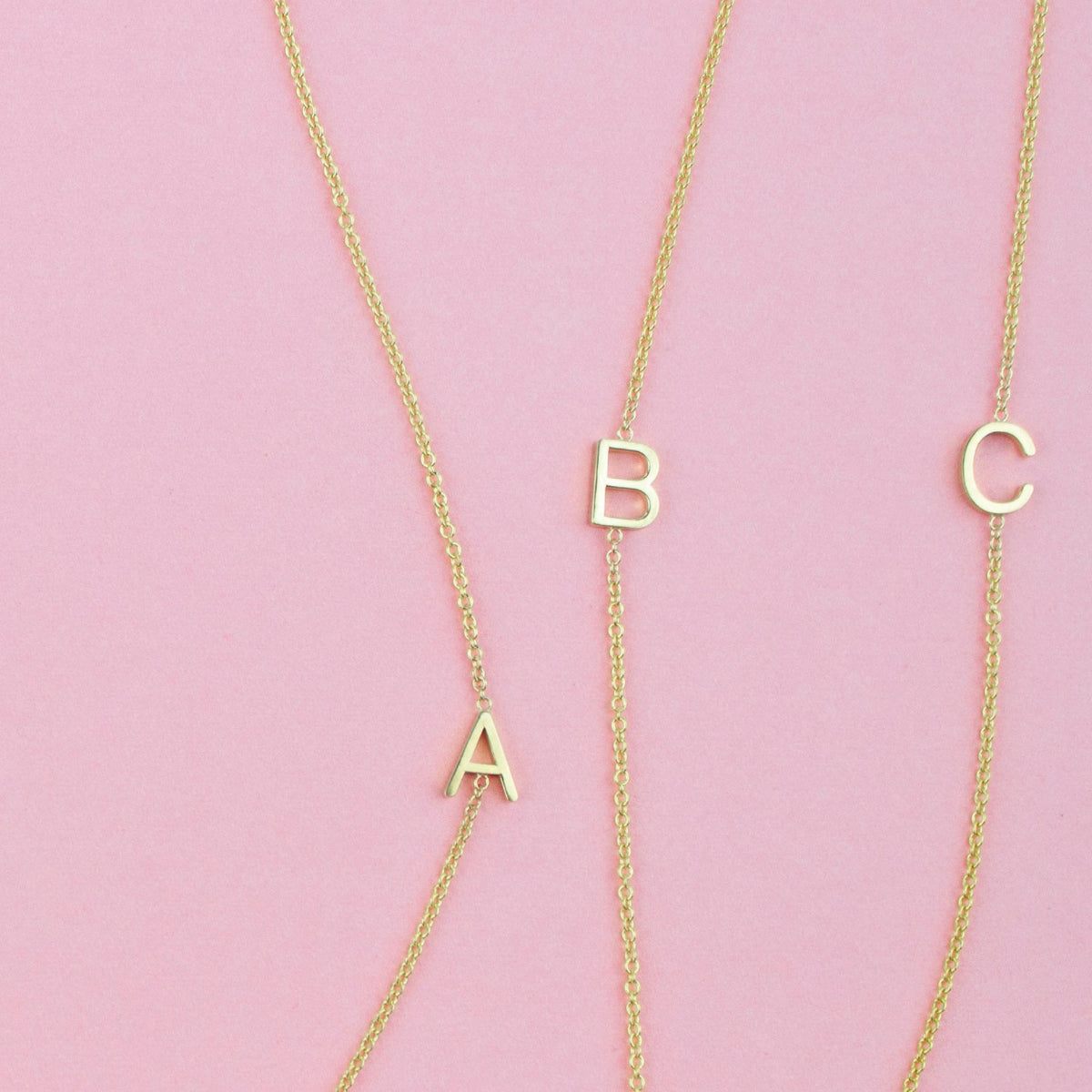 Three gold necklaces with single letter charms A, B and C on a pink background. 