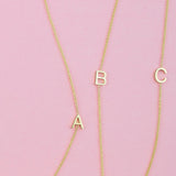 Three gold necklaces with single letter charms A, B and C on a pink background. 