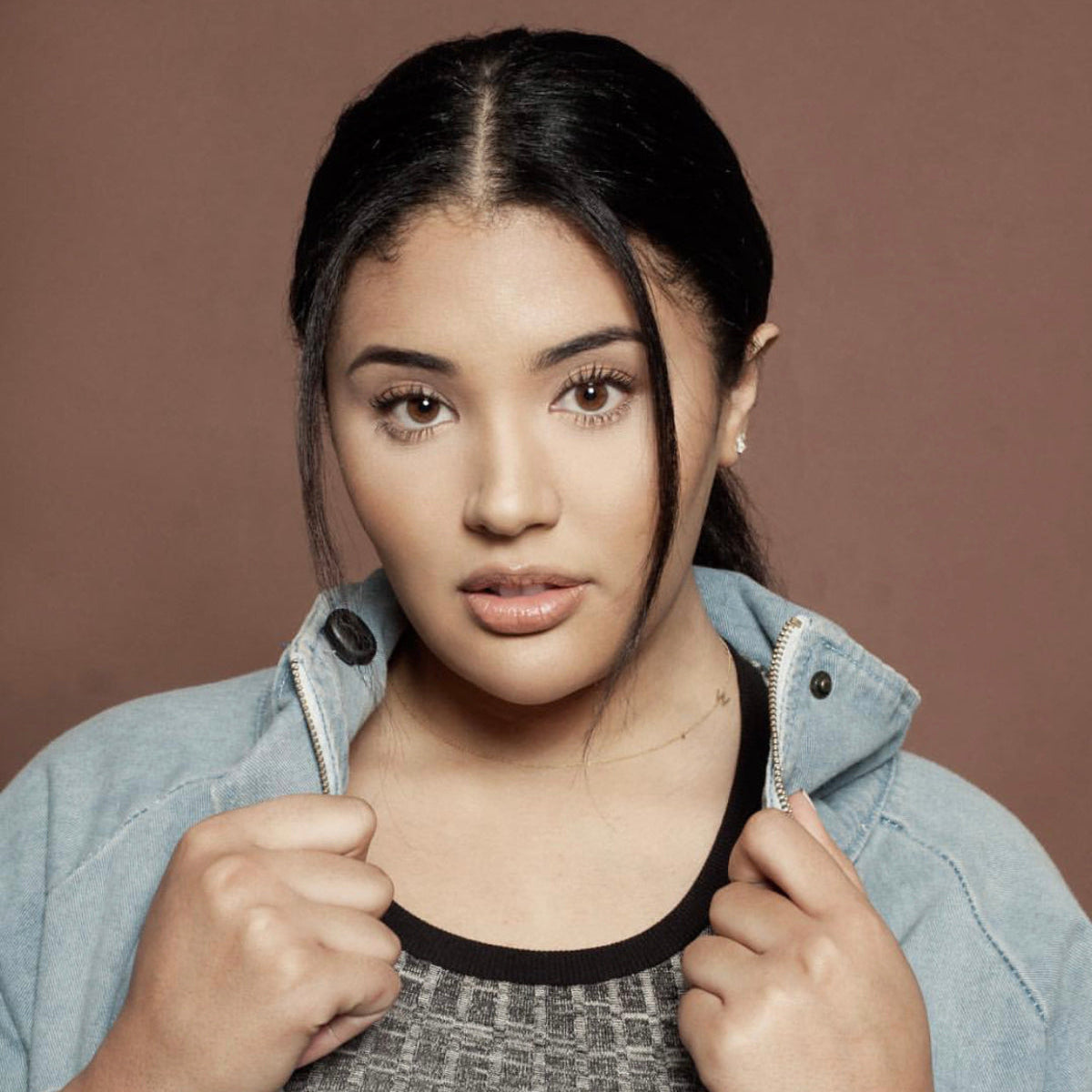 A woman wearing a dark grey top and denim jacket, a necklace with a letter W charm and stud earrings.
