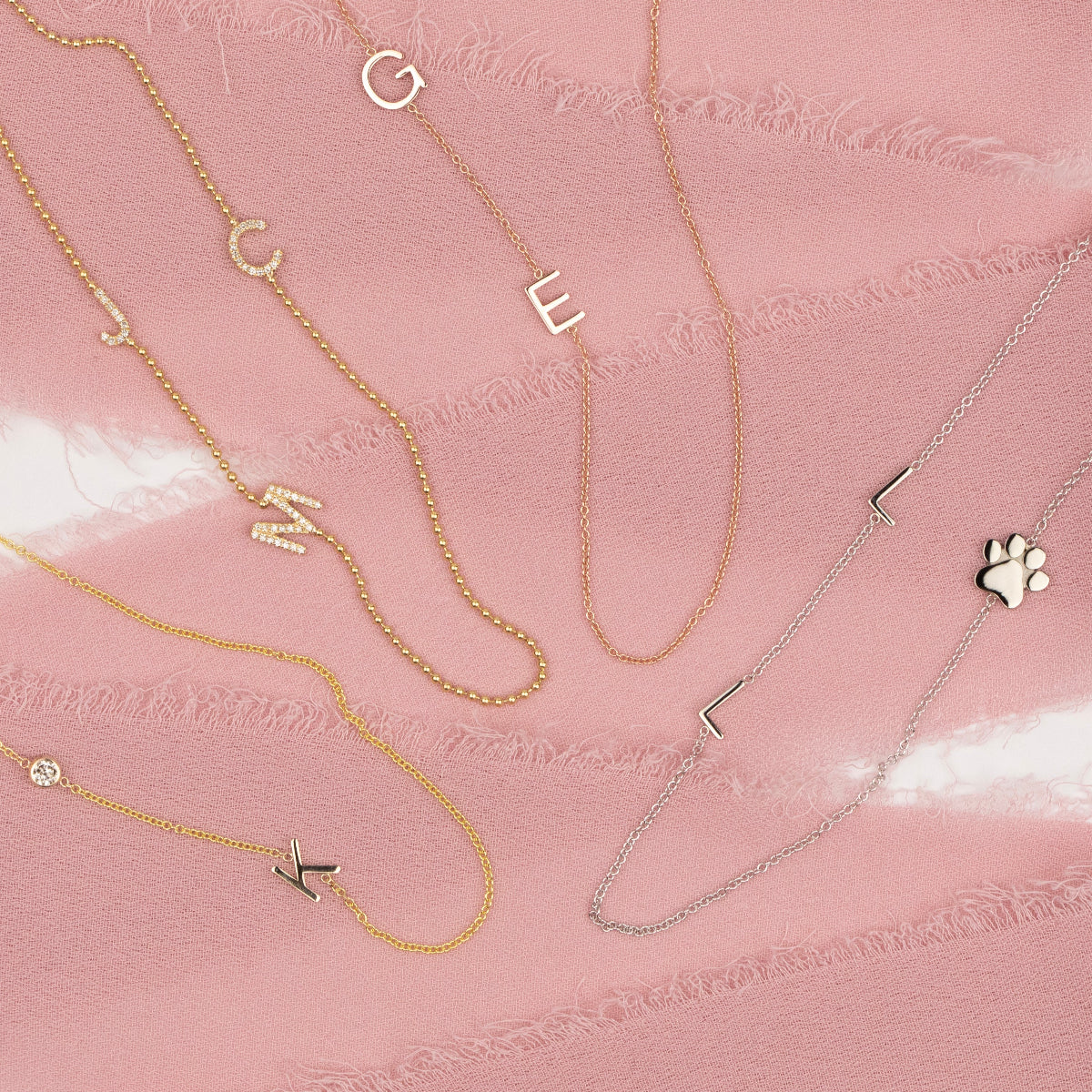 Four Custom Gold Necklaces: a yellow gold necklace with a K letter and birthstone charm, a yellow gold beaded necklace with pavé letter charms J, M and C, a rose gold necklace with two letter charms G and E, and a white gold necklace with two L letter charms and a paw, on a pink background.