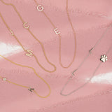 Four Custom Gold Necklaces: a yellow gold necklace with a K letter and birthstone charm, a yellow gold beaded necklace with pavé letter charms J, M and C, a rose gold necklace with two letter charms G and E, and a white gold necklace with two L letter charms and a paw, on a pink background.