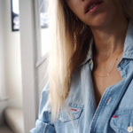 A close-up of a woman's neck and chest area wearing a gold necklace with a letter M charm and a denim shirt.