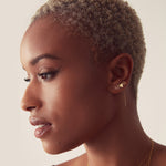 A woman wearing a 14K Gold Pave Letter Stud in M, a Charm Stud - Heart and a 14k Gold Letter Threader Earring in B against a light background.