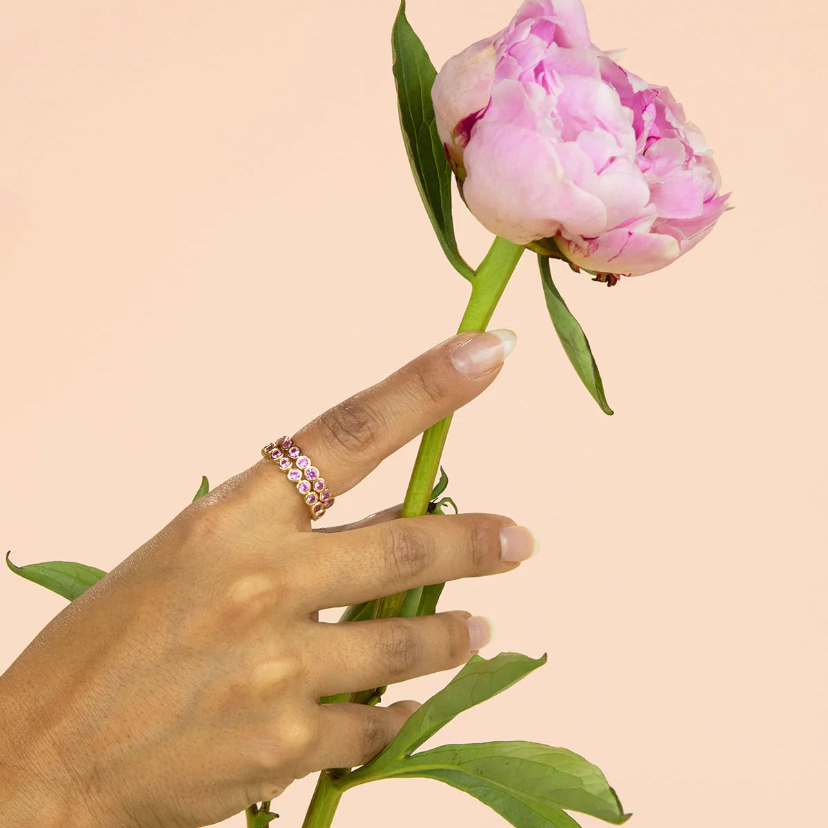 A hand wearing two The Peony Bouquet Rings holding a peony flower against a light peach background. 