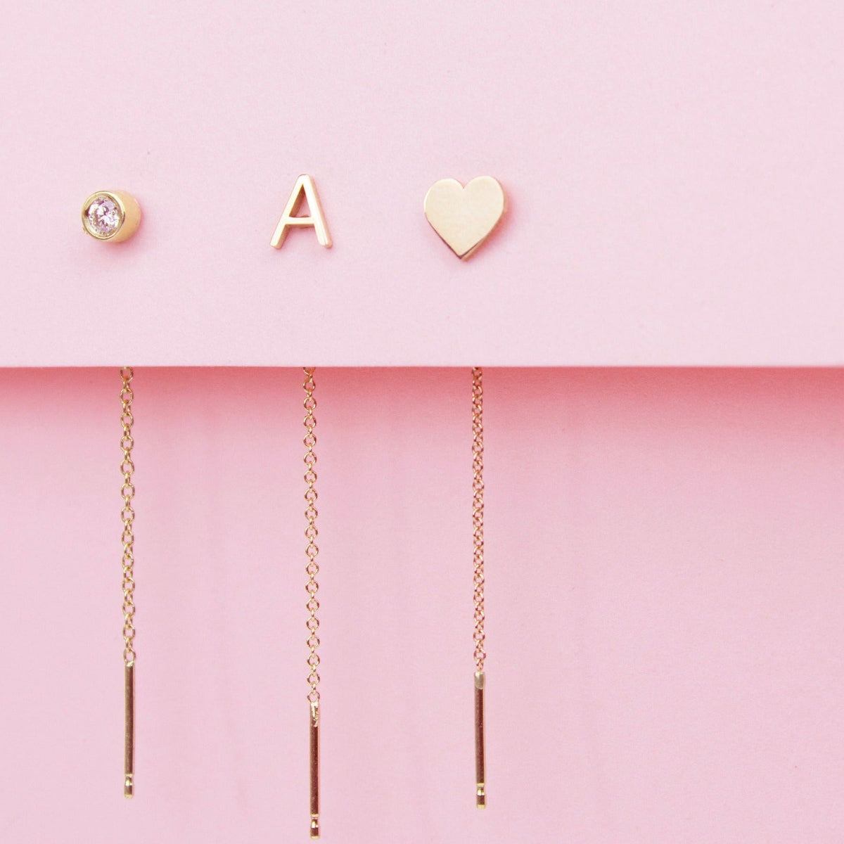 Three gold threader earrings - one with a diamond birthstone, one with a letter A stud and one with a heart stud, against a light pink background. 