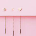 Three gold threader earrings - one with a diamond birthstone, one with a letter A stud and one with a heart stud, against a light pink background. 