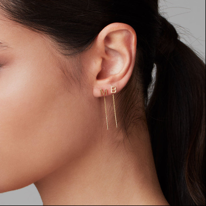 A close-up of a woman's side profile wearing two yellow gold threader earrings with letter studs M and B.