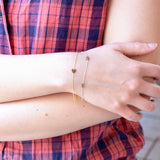 Close-up of a person's wrist wearing two Custom Gold Bracelets - one yellow gold with a heart charm and one white gold with two letter E charms, with a plaid shirt. 