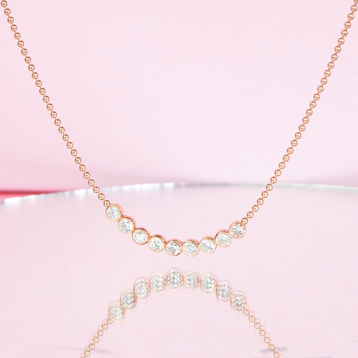 A Diamond Arc Layering Necklace in yellow gold against a light pink background. 