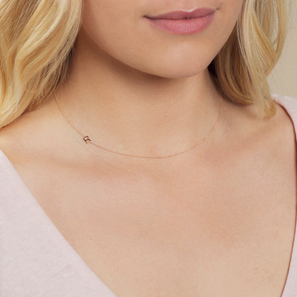 A close-up of a woman's neck wearing a rose gold necklace with a letter R charm. 
