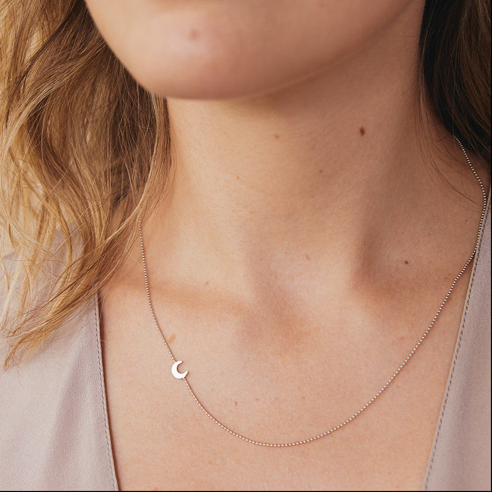 Close-up of a woman's neck wearing a white gold 14k Gold Asymmetrical Charm Necklace - Moon with a bead chain, styled  in a neutral top against a beige background.