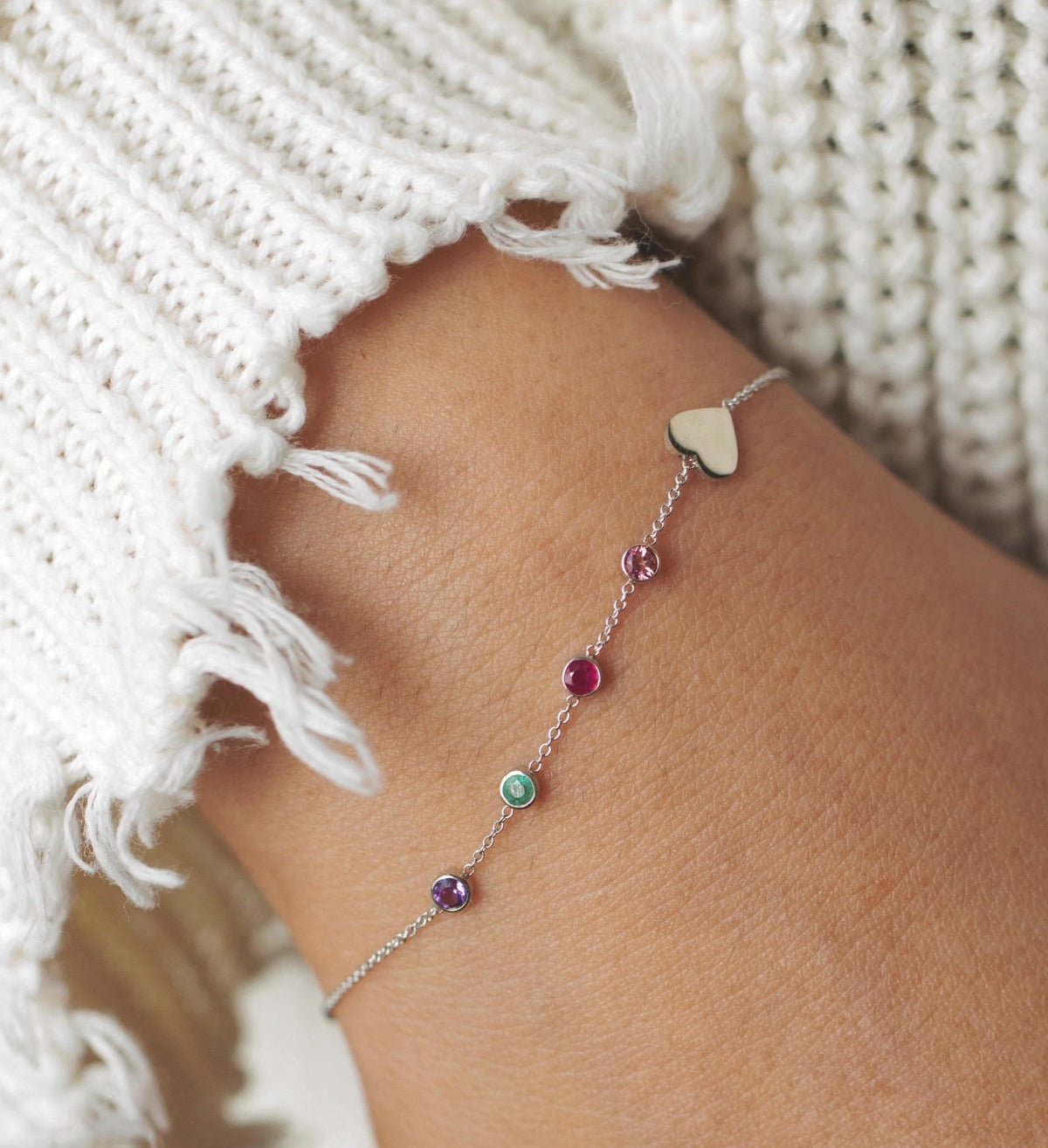 A close-up of a wrist wearing a Custom Gold Bracelet – 5 Letters – with four birthstones and one heart charm, styled with a white knit.