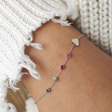 A close-up of a wrist wearing a Custom Gold Bracelet – 5 Letters – with four birthstones and one heart charm, styled with a white knit.