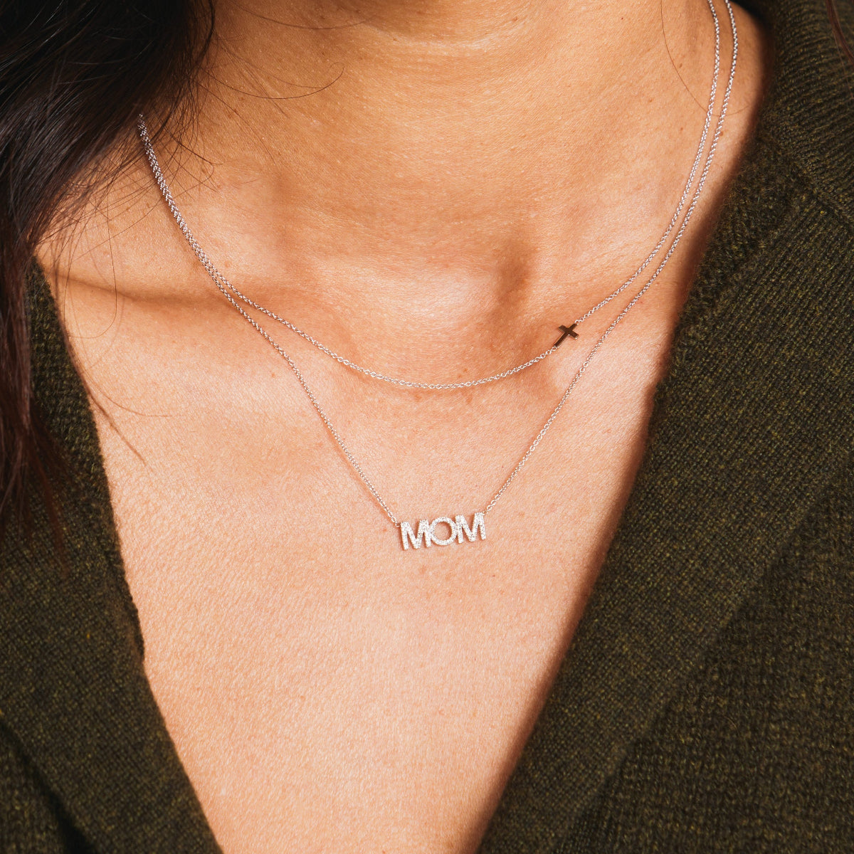 A close-up of a woman's neck wearing 14k Gold Asymmetrical Charm Necklace - Cross and a Pavé MOM Necklace, both in white gold, styled with a brown knit.