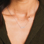 A close-up of a woman's neck wearing 14k Gold Asymmetrical Charm Necklace - Cross and a Pavé MOM Necklace, both in white gold, styled with a brown knit.