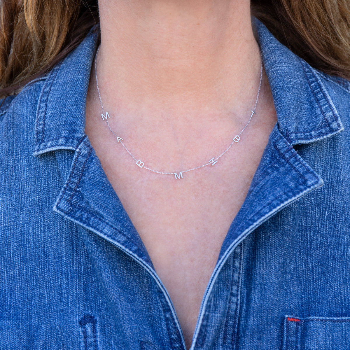 A close-up of a woman's neck wearing a Custom Gold Necklace in white gold with seven letter charms (M, A, B, M, H, B and T) and a blue denim shirt. 